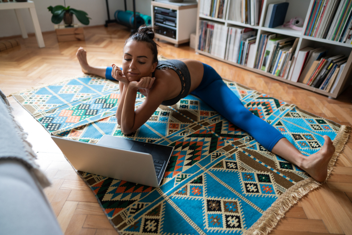 Eine Frau in Yogaleggins macht in ihrem WOhnzimmer auf einem Teppich einen Spargart und schaut etwas in Ihrem Laptop an.